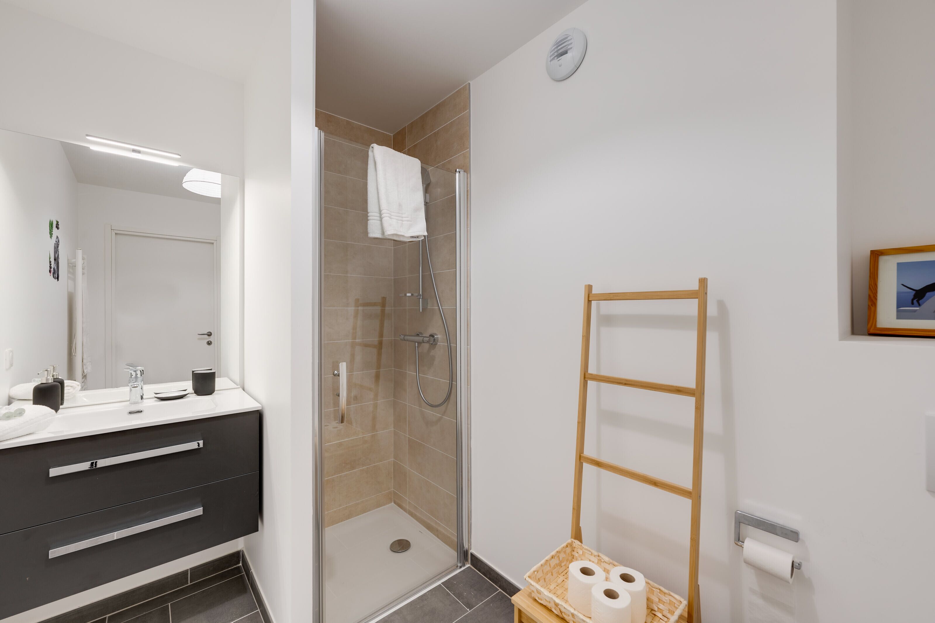 A modern bathroom with a glass-enclosed shower, a white sink with a black cabinet, and a wooden ladder leaning against the wall.