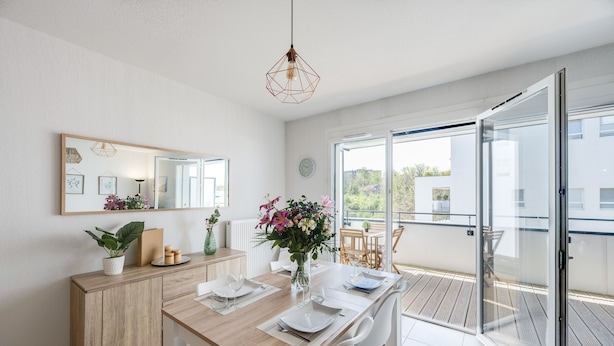 Un espace repas moderne avec une table en bois, des chaises blanches et une porte vitrée donnant sur un balcon.