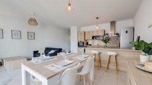 Une cuisine moderne avec des meubles en bois, un coin repas doté d’une table et de chaises blanches, et des luminaires suspendus.