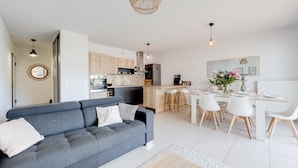 Un salon moderne avec un canapé gris, un coin repas avec des chaises blanches, une cuisine avec des meubles en bois et des luminaires suspendus.