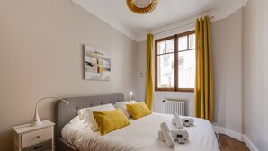 A neatly made bed with white linens and two yellow pillows, a bedside table with a lamp, and a window with yellow curtains.