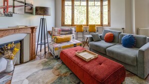 A cozy living room with a fireplace, a large window with a view of trees, a grey sofa with colorful cushions, and a red ottoman.