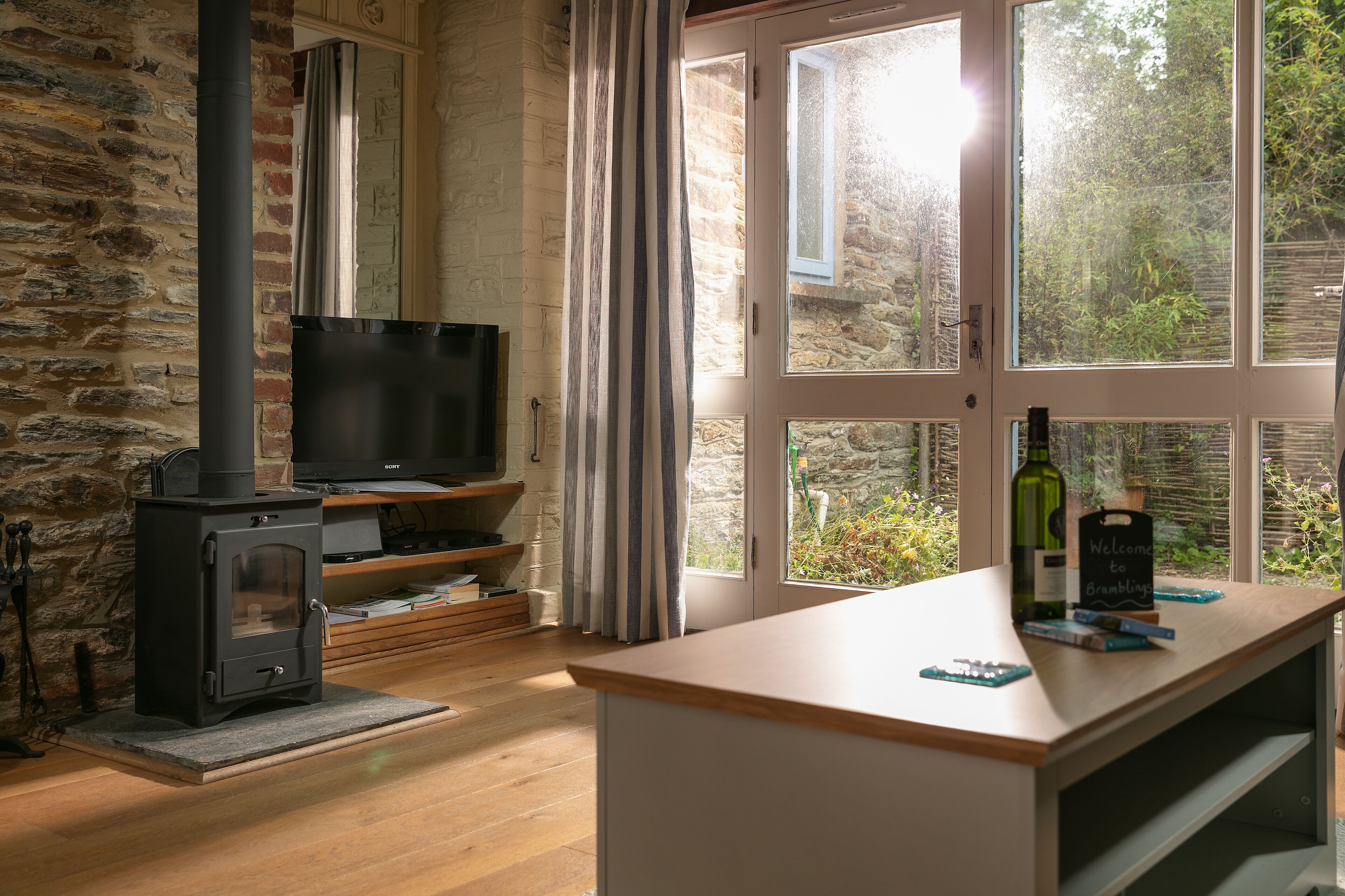Ein gemütliches Wohnzimmer mit einer Steinwand, einem Kamin, einem Fernseher und einem großen Fenster mit Blick auf Grünflächen.
