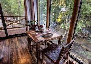 A wooden dining table with two chairs, a glass of water, and a plate set for two, overlooking a forested area with a stream.