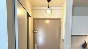 Una camera d'albergo con pavimento in legno, un letto con biancheria bianca, una porta con maniglia, un cestino nero e un angolo cottura con fornello e forno.