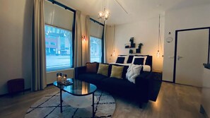 A modern living room with a black sofa, a glass coffee table, and a large window with curtains. - Marble Times (Groningen)