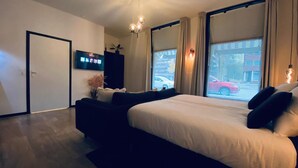 Habitación de hotel moderna con una cama grande, un sofá oscuro, un televisor de pantalla plana y una ventana con vistas a la calle.