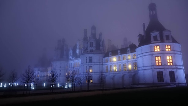 Un edificio grande e histórico con múltiples torres y ventanas iluminadas, rodeado de árboles desnudados y luces navideñas.