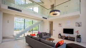 Un vaste espace de vie comprenant un grand canapé gris, une table basse et un téléviseur posé sur un meuble en bois. Un escalier avec des rampes en verre mène à un niveau supérieur.