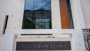 Un bâtiment moderne doté d’une grande baie vitrée qui reflète un paysage montagneux. Le nom « VILLA BARCELONA » est affiché en caractères bien visibles sur la façade.