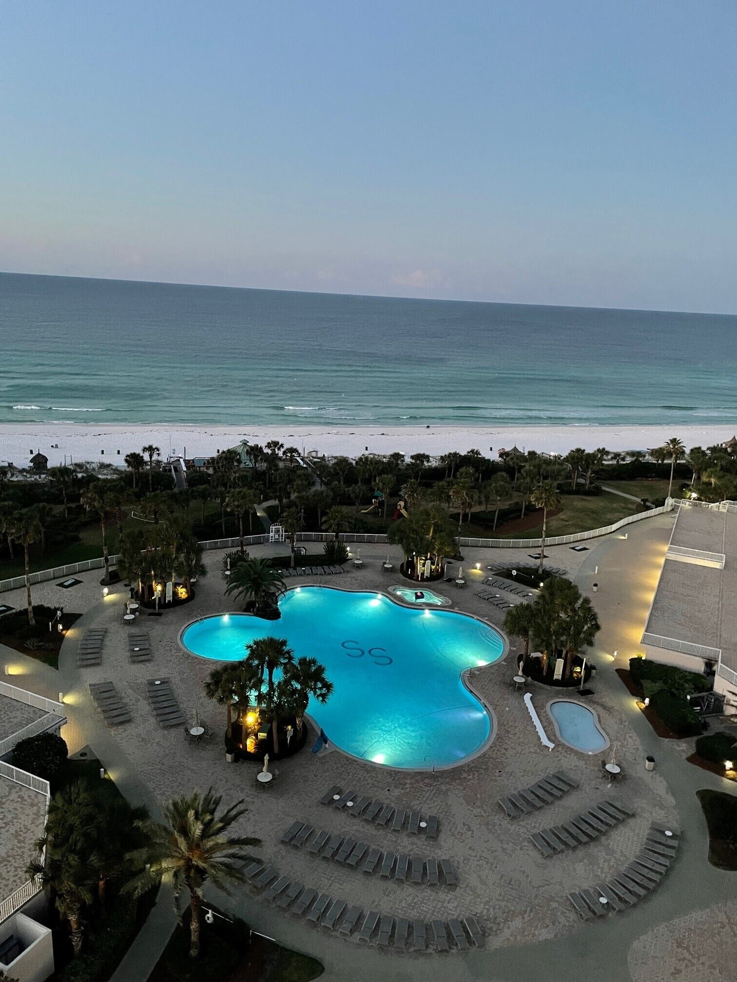 A resort with a large swimming pool surrounded by lounge chairs and palm trees, overlooking a beach and the ocean.
