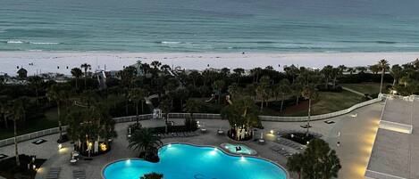 A resort with a large swimming pool surrounded by lounge chairs and palm trees, overlooking a beach and the ocean.