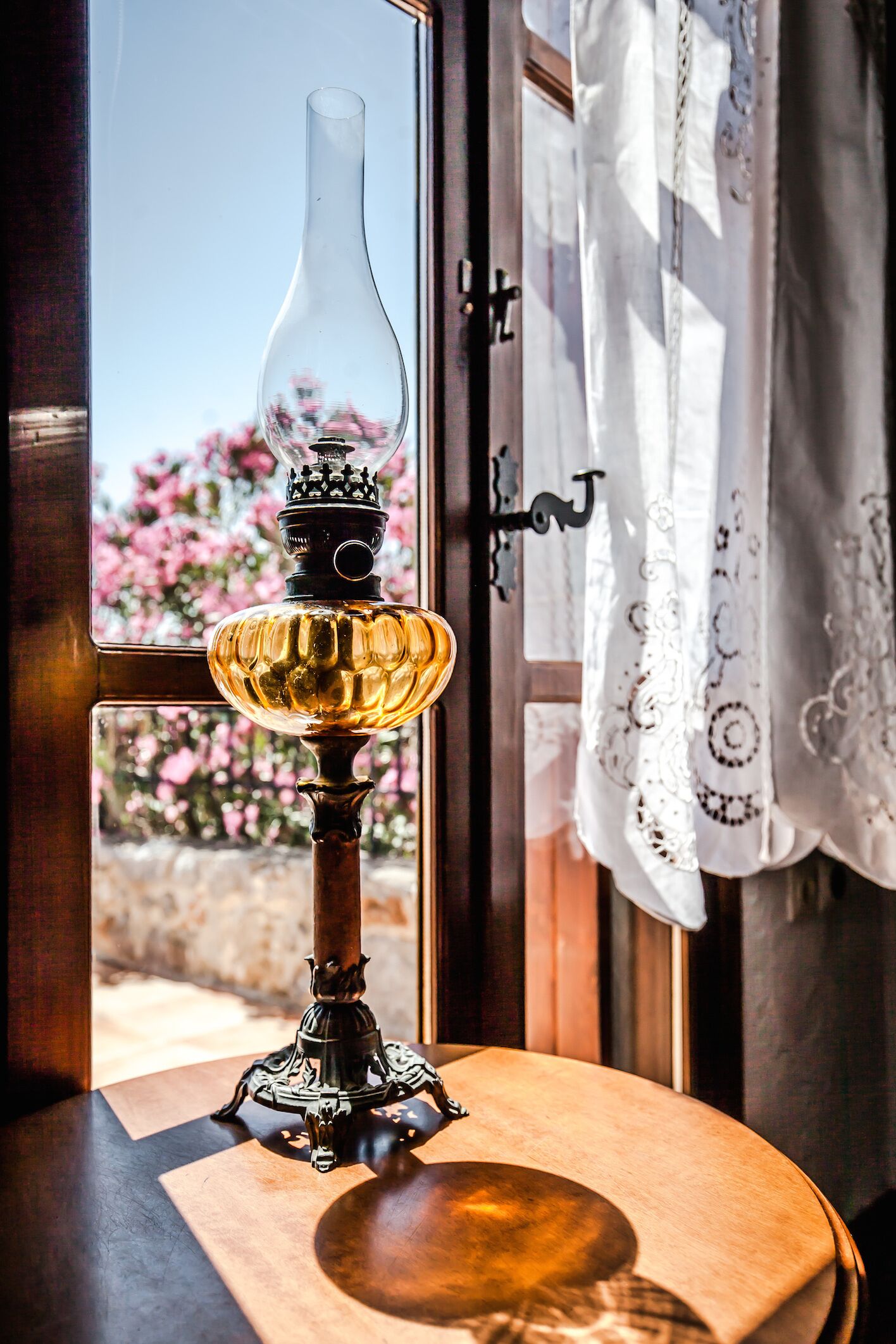 Una lámpara de mesa con pantalla de vidrio transparente y base decorativa, sobre una mesa de madera cerca de una ventana que da a flores rosadas y un muro de piedra.