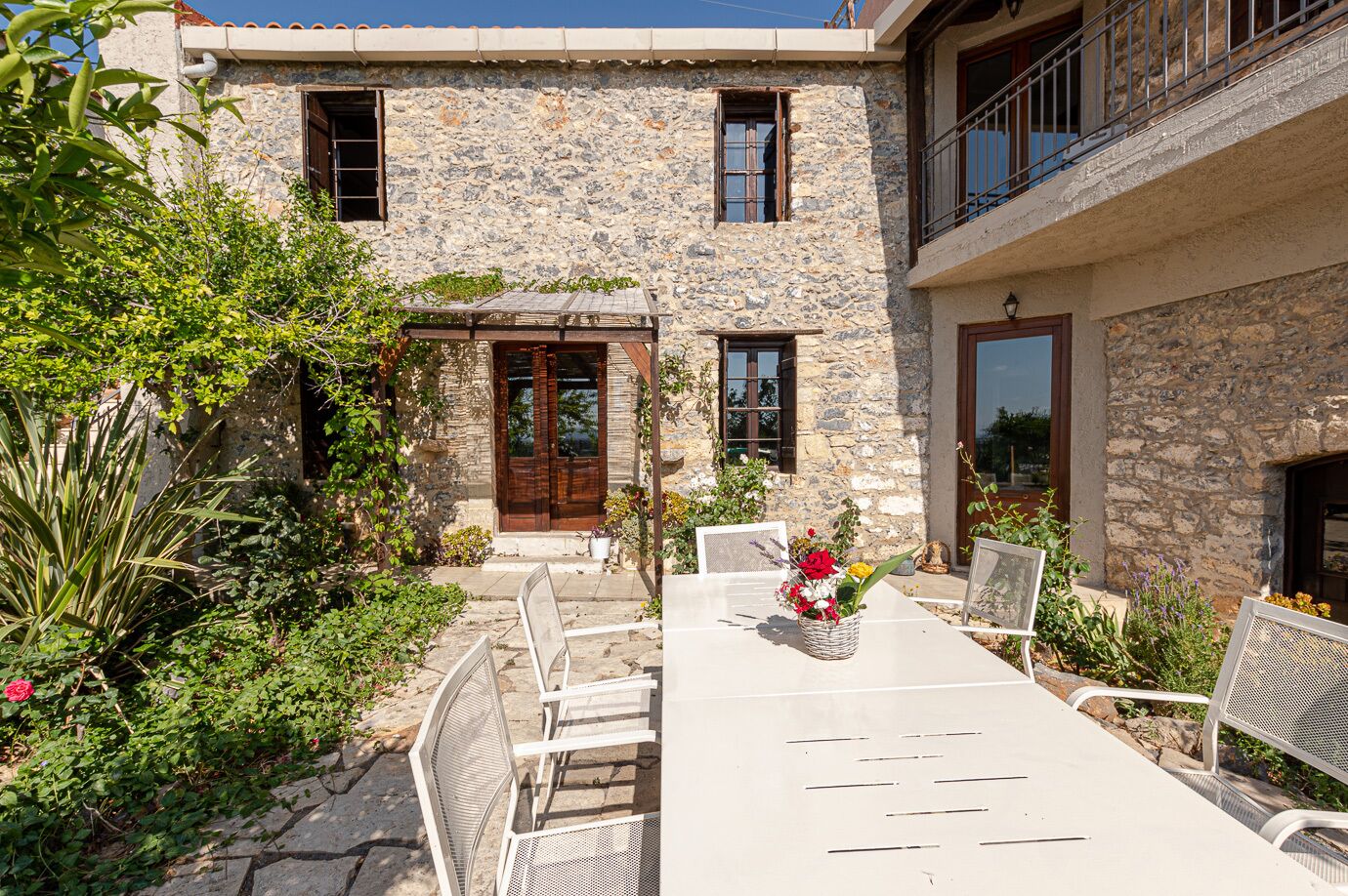 A stone building with a balcony, a wooden door, and a table with chairs and a vase of flowers.