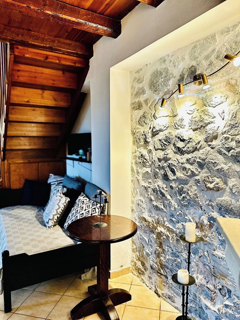 A bedroom with a wooden ceiling, a stone wall, a bed with patterned pillows, a small round table, and a floor lamp.