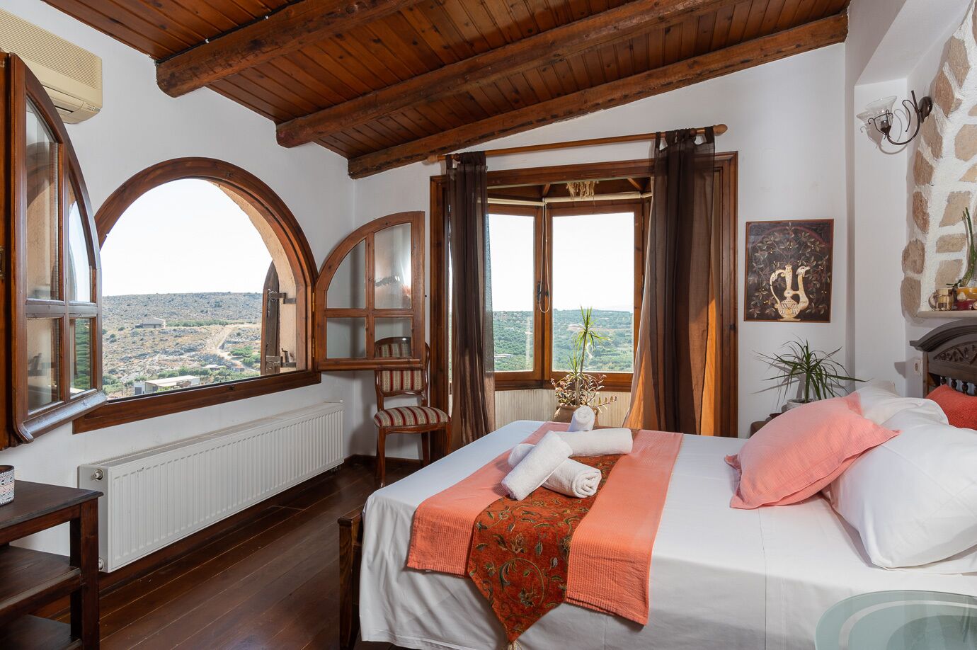A bedroom with a large bed, a chair, a table, and a view of the landscape through the windows.