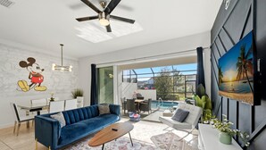 Uma sala de estar moderna com um sofá azul, uma mesa de centro e um tapete estampado. Há uma área de jantar com uma mesa e cadeiras brancas, e uma janela grande que oferece vista para o exterior.