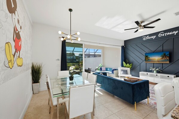A modern living room with a glass dining table, white chairs, a blue sofa, and a ceiling fan.