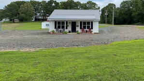 A small white building with a metal roof, a front porch with two chairs, and a gravel driveway. - Cozy, Modern Cabin with Private Hot Tub, Sleeps 6, Direct ATV Trail Access! (Hatfield)