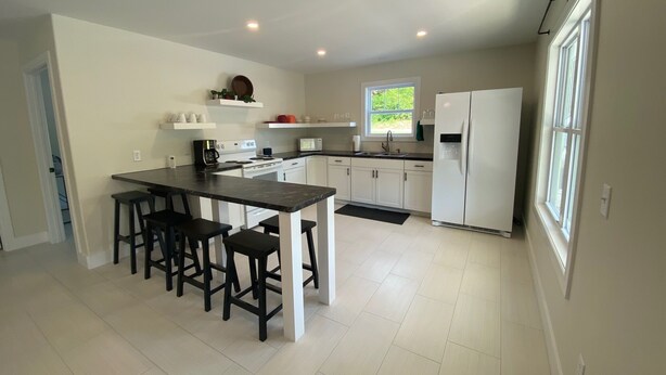 A modern kitchen with white cabinets, a central island with bar stools, a refrigerator, and a window providing natural light. - Cozy, Modern Cabin with Private Hot Tub, Sleeps 6, Direct ATV Trail Access! (Hatfield)
