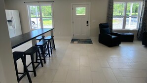 A spacious living area with a dining table and chairs, a black sofa, and a large window with a view of greenery. - Cozy, Modern Cabin with Private Hot Tub, Sleeps 6, Direct ATV Trail Access! (Hatfield)