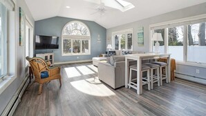 A spacious living room with a large window, a ceiling fan, and a dining area with stools. - Poppy’s Hampton House | 3BR + Bonus | Near Beaches & LIRR (Hampton Bays)