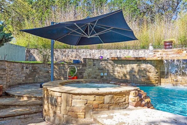 A stone hot tub with a waterfall feature, surrounded by a stone wall and a large umbrella.