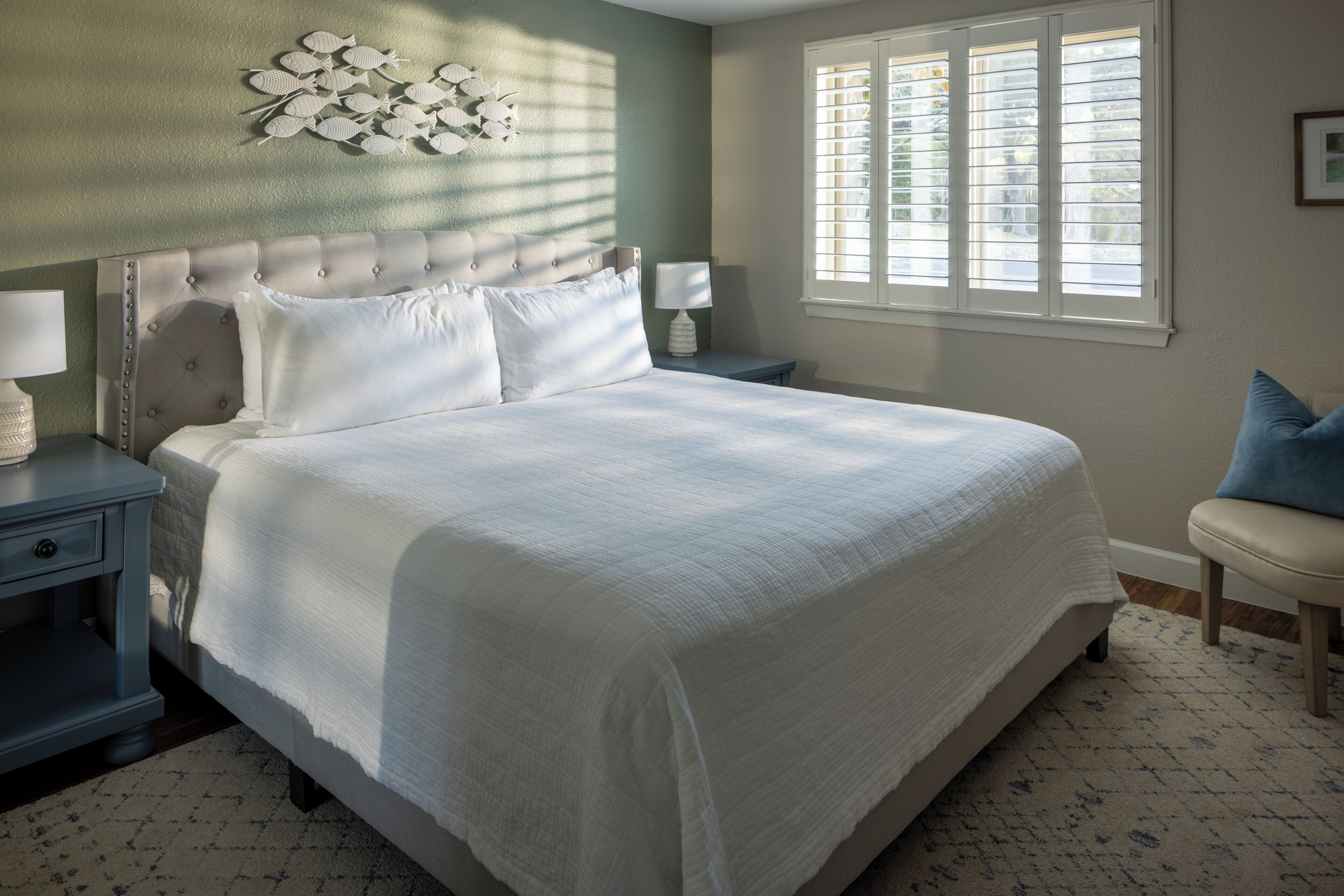 A bedroom with a large bed, two bedside tables, a nightstand, a window with blinds, and a wall-mounted light fixture.