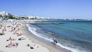 A crowded beach with people sunbathing and swimming, a pier extending into the water, and buildings in the background.