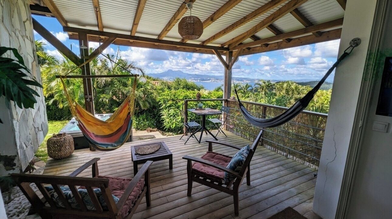 A wooden deck with a hammock, chairs, and a table overlooking a lush landscape with mountains in the distance.