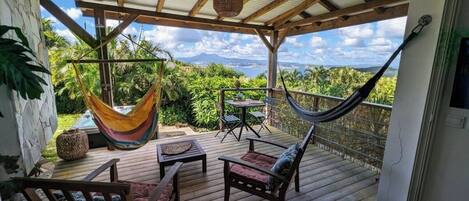 Terraza de madera con una hamaca, sillas y una mesa, con vistas a un paisaje exuberante y montañas a lo lejos.