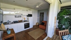 Een compacte keuken met witte kastjes, een magnetron en een fornuis. Er staat een houten tafel met een blauwe kom en een rieten mand. Op de vloer ligt een beige vloerkleed, en er is een houten bank bij de deur.