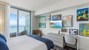 A hotel room with a bed, a desk, a chair, a large window with a view of the ocean, and various artworks on the wall.
