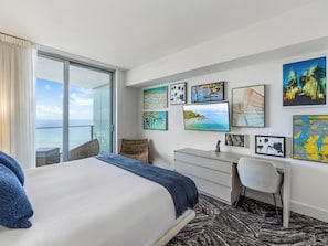 Quarto de hotel com uma cama, uma escrivaninha, uma cadeira, uma janela ampla com vista para o oceano e diversas obras de arte nas paredes.