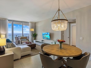 A modern living room with a round dining table, a sofa, a television, and a large window with a view of the ocean.