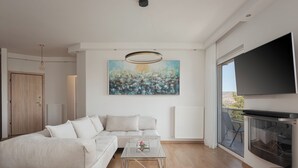 A modern living room with a sectional sofa, a glass coffee table, a painting on the wall, a TV, and a balcony with outdoor furniture. - Athenian Riviera Sea View Penthouse (Vari-Voula-Vouliagmeni)