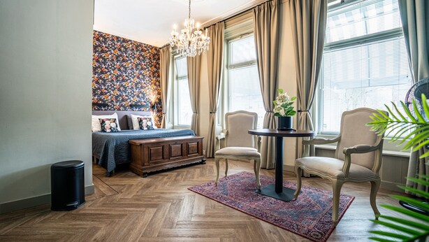 A room with a wooden herringbone floor, a bed with floral bedding, a bench, a round table with a chair, and a chandelier. - The Hogg House (Groningen)