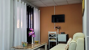Ein Zimmer mit einer Couch, einem kleinen Tisch mit Blumen in einer Vase, einem an der Wand montierten Fernseher und einem Schreibtisch mit Stuhl.