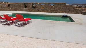 A pool with red chairs, a stone wall, and a clear blue sky. - NikoRamaVilla  Swimming pool and BBQ with sea and mountains view (PAROS)