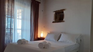 A neatly made bed with rolled towels, a bedside lamp, and a window with curtains. - NikoRamaVilla  Swimming pool and BBQ with sea and mountains view (PAROS)