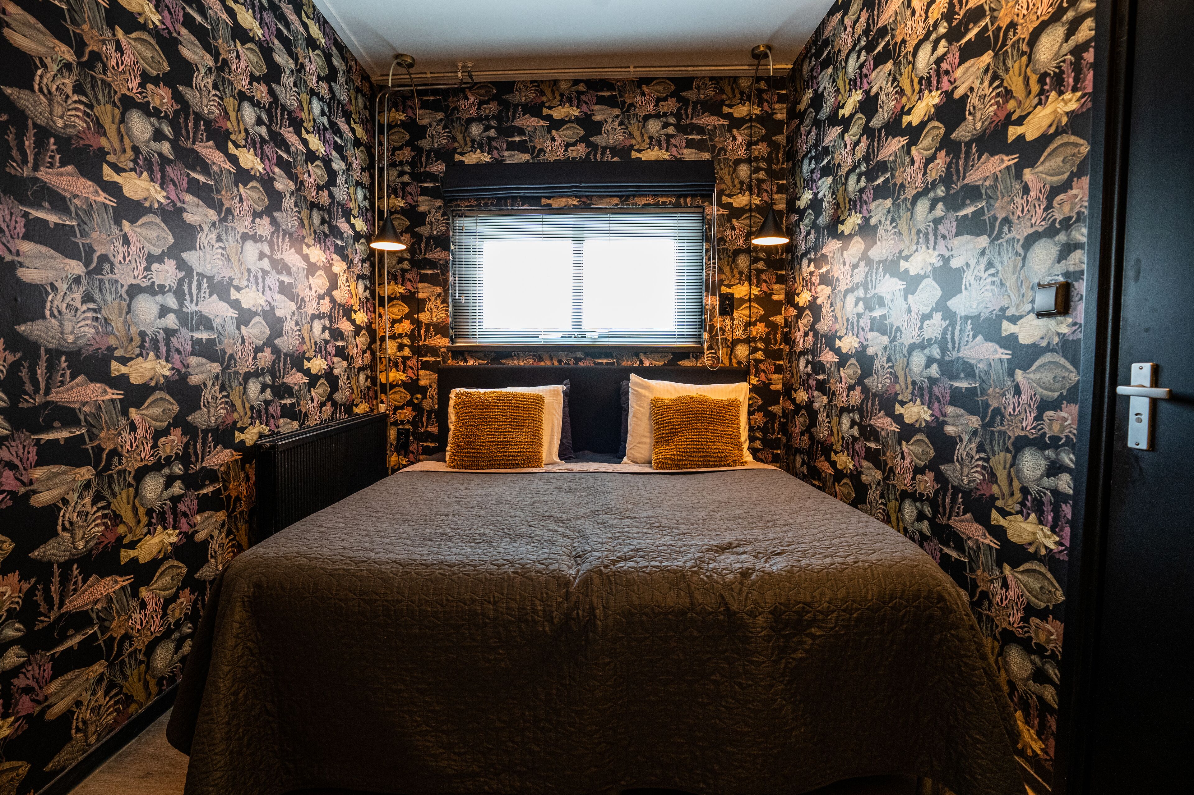 A bedroom with a bed centered between two walls covered in marine life wallpaper, a window with blinds, and a radiator below the window.