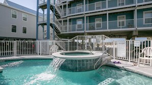 Une piscine avec un jacuzzi, entourée d’une clôture blanche, et un bâtiment avec des balcons en arrière-plan.