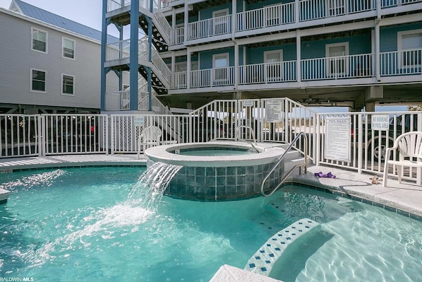 A swimming pool with a hot tub, surrounded by a white fence, and a building with balconies in the background. - Newly Updated Beach Front Retreat (Gulf Shores)
