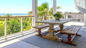 Área externa coberta com mesa e bancos de madeira, com vista para uma praia e palmeiras.