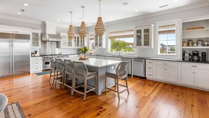 Cozinha moderna com armários brancos, uma ilha grande com banquetas, eletrodomésticos de aço inoxidável e iluminação pendente.