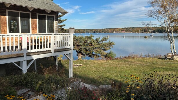 A wooden cabin with a deck overlooking a lake, surrounded by greenery and flowers. - Cozy, ocean-side retreat (Sedgwick)