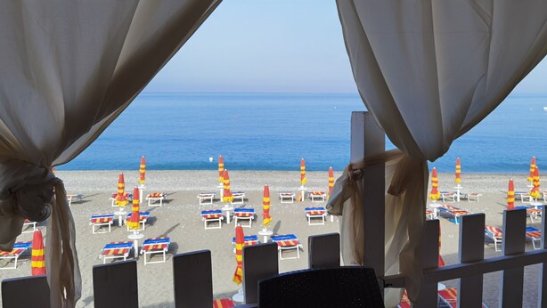 Vista sulla spiaggia da un balcone con ringhiera, con sedie a sdraio, ombrelloni e barche in acqua.
