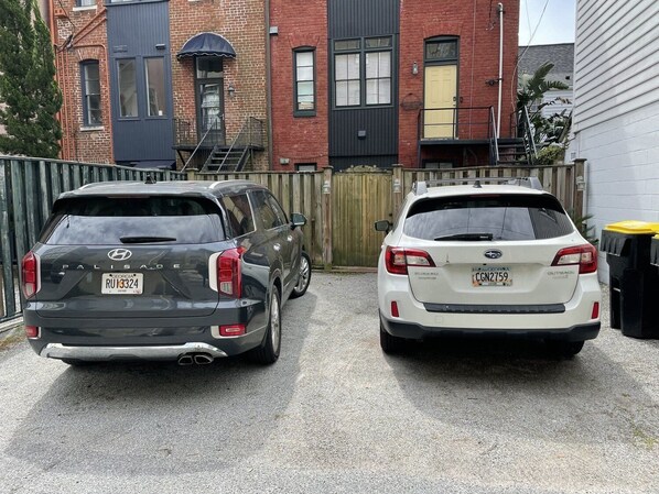Dos coches aparcados en una entrada con un edificio de ladrillo y una valla de madera al fondo.
