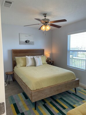 A bedroom with a wooden bed, a ceiling fan, a window with blinds, and a colorful striped rug.
