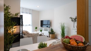 A modern living room with a flat-screen TV, white furniture, a dining area with a wooden table, and a fruit basket.